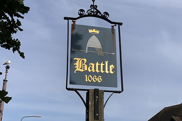 A town sign hanging within a rought iron frame atop of a tall post. The rectangular sign includes a 1066 battle helmet above the word "Battle 1066" in gold on a grey background